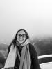Black and white photo of a white woman wearing glasses and a scarf around her neck, smiling, in front of fog