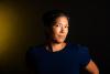 Photograph of Alexandra Hui in a dark blue shirt against a dark background. Her face is turned away from the camera but her eyes are looking at the camera. Her face and arm and some of the wall behind her is illuminated in a golden yellow.