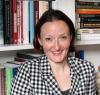 A smiling woman with a checked blazer and short brown hair stands in front of a bookcase with books on it.